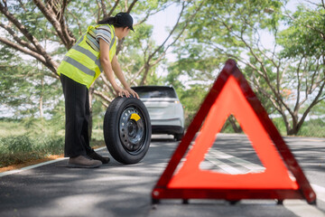 Person holds a spare tire on a road with trees and a car in the background during daytime © 2B
