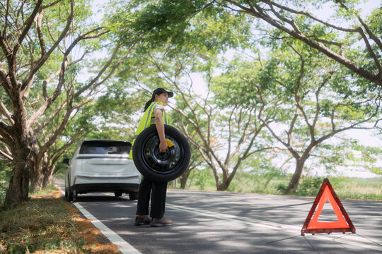 Worker changes tire on roadside near car with hazard sign during a sunny day in a wooded area