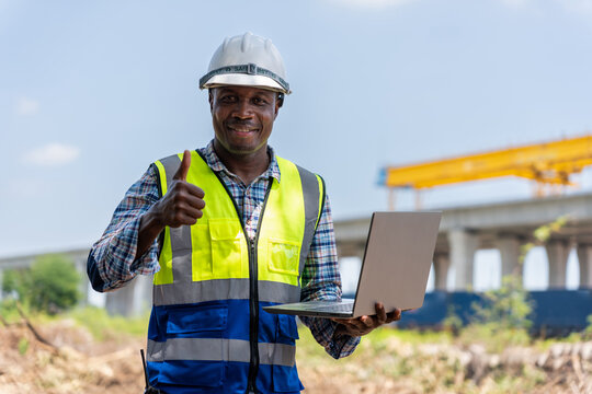 Happy engineer holding laptop and giving thumbs up at construction site, Professional supervisor successful with project at industrial plant, Smiling technician showing approval at infrastructure site