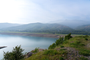 Fototapeta premium Beautiful vista of Charvak reservoir with lush green hillsides and misty mountains in the distance, Uzbekistan. Concept: Central Asian ecotourism