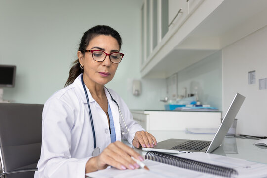 Serious senior medical professional woman in glasses and lab coat reviewing medical records in paper notebook, reading patients history, research results, using laptop at workplace