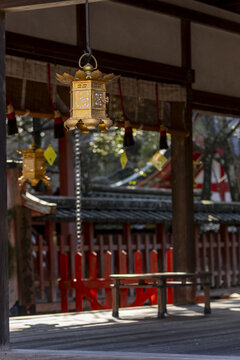 View of an ornate golden hanging lantern under the eaves of a traditional wooden temple building with red fences in the background in Nara, Nara, Japan.