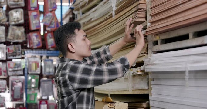 Asian adult man working as woodworking business owner reaches wooden planks from stacked shelves inside local hardware store wood section while checking stock inventory quality for interior furniture