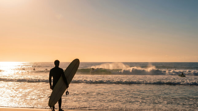 Male surfer in black wetsuit, with long surfboard held underarm, standing on sandy shore looking out at sea waves, ocean beach during golden sunset, sports lifestyle photography