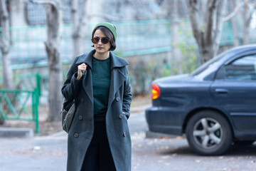 Fototapeta premium Portrait of a woman wearing a dark coat, green beanie, and sunglasses standing outdoors with a car in the blurred background.