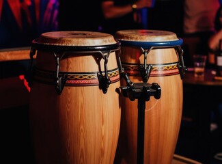 Fototapeta premium Pair of conga drums with detailed wood grain sit ready for music performance. Traditional percussion instruments await rhythm in dimly lit bar setting. Colorful band adorns drums for vibrant salsa