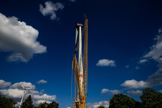 View of wind turbine construction with a tall yellow crane assembling a large turbine under a blue sky with white clouds Germany.