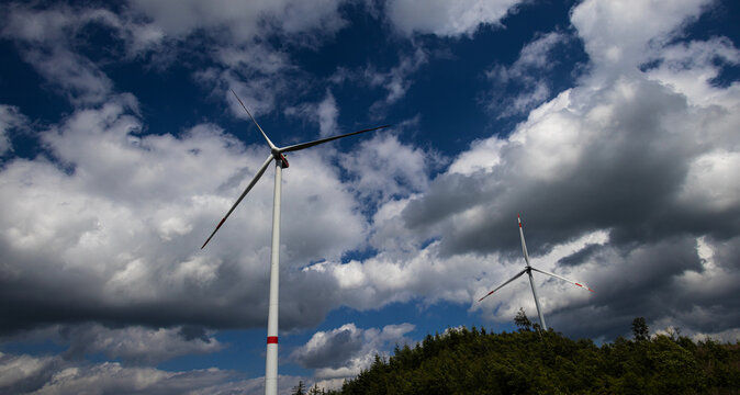 View of two modern wind turbines standing on a lush green forested hill under a sky filled with heavy white and dark clouds in Germany.