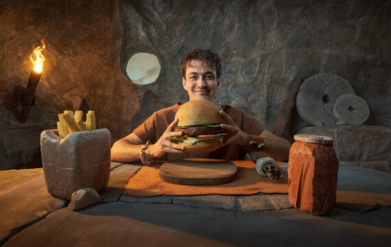 Funny Smiling Young Prehistoric Caveman Holding Stone Hamburger Fast Food in a Cave