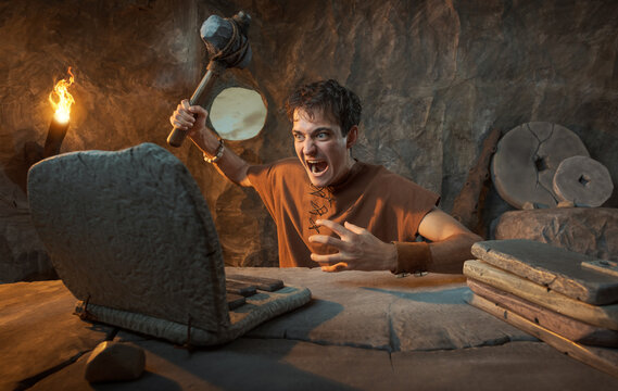 Funny Angry Young Prehistoric Caveman Smashing Stone Laptop with Hammer in a Cave