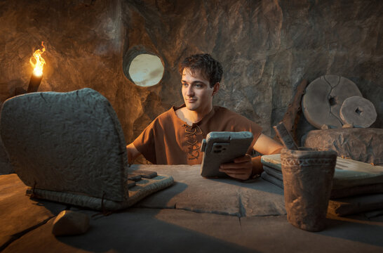 Funny Young Prehistoric Caveman Working with Stone Laptop and Smartphone in a Cave