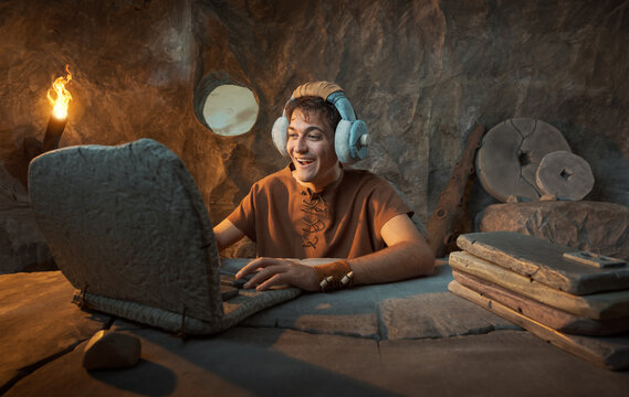 Funny Happy Young Prehistoric Caveman with Stone Headphones Using Laptop in a Cave