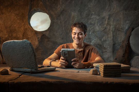 Funny Smiling Young Prehistoric Caveman Distracted by Stone Smartphone in a Cave