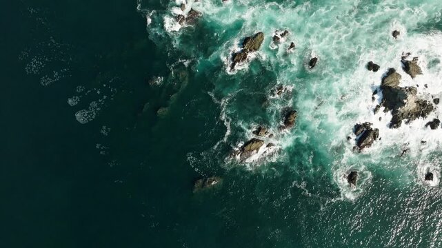 Aerial backdrop of turquoise ocean waves breaking around offshore rocks along the coast, drone establishing background, top down