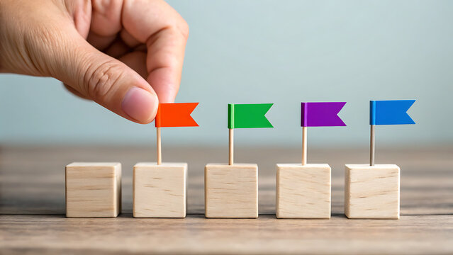 A hand placing a small orange flag on a wooden block in a row of five blocks with different colored flags representing goals and milestones