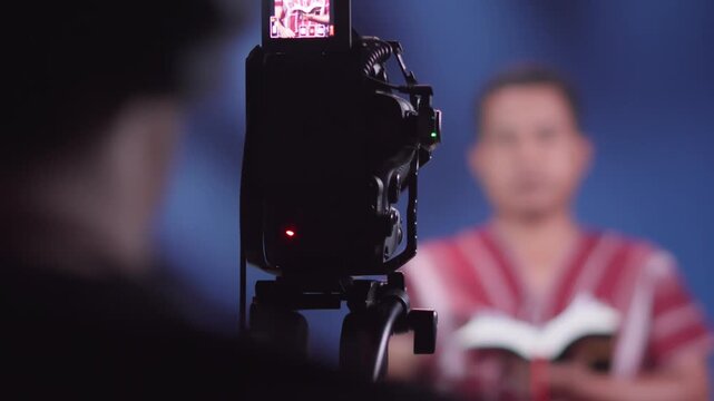 "Close up focus of modern professional camera filming Southeast Asian ethnic minority man vlogger sharing encouraging Bible verses, indigenous Christian content creator talking to viewers, recording f