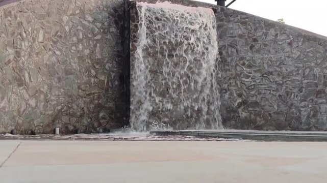 Stone wall channels flowing water into a pool below. Water cascades down rugged, textured surface. Scene suggests tranquil, natural design in urban setting