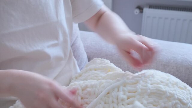 Female hands knitting a cozy white blanket using super chunky plush yarn at home.