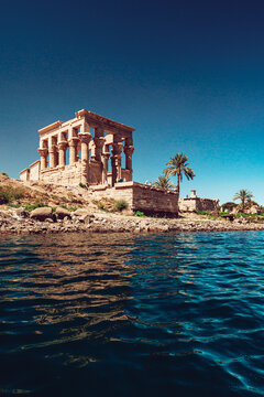 View of the ancient Trajan's Kiosk at Philae Temple with stone columns and palm trees under a clear blue sky from the Nile river in Edfu, Aswan Governorate, Egypt.