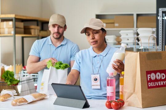 Young delivery workers preparing grocery pickup order and checking tablet at fulfillment point. Concept for food delivery service, retail logistics, online shopping marketing