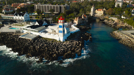 White and blue striped lighthouse stands on rugged coastal rocks next to buildings and serene bay waters in Cascais, Portugal, offering a beautiful travel destination © Dark Artistic