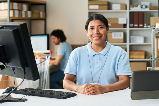 Portrait of young Latina woman managing order pickup desk in delivery warehouse while coworker processing shipments in background. Useful for e commerce logistics marketing