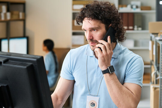 Medium shot of young man managing order pickup point, speaking on smartphone and checking monitor. Useful for delivery service, retail logistics, fulfillment
