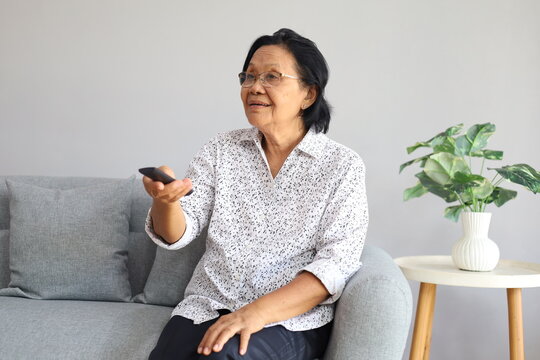 Happy smiling Asian senior woman sitting on couch in the living room, pointing tv remote controller to switch channels, watching favorite movie online series