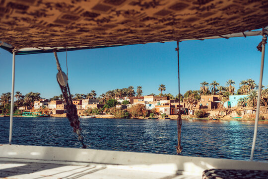 View of the Nile River bank with traditional village houses and palm trees seen from a boat in Edfu, Aswan Governorate, Egypt.