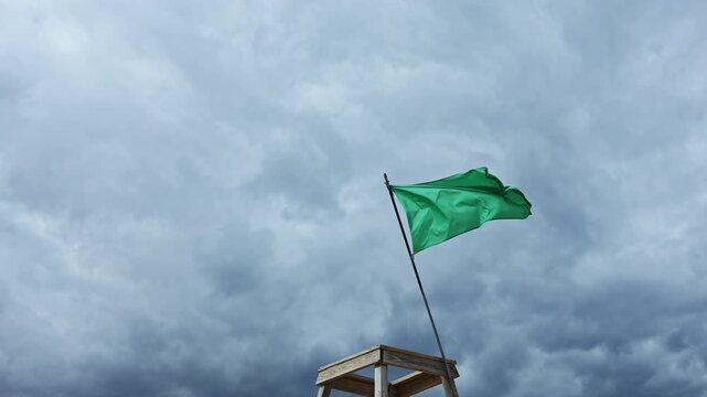 The green flag stands on a wooden post, fluttering in the wind. Dark clouds fill the sky, indicating potential rain. The scene shows an outdoor area with changing weather.