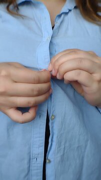 Close-up of a plus-size woman's hands buttoning up a light blue cotton shirt over a black top. Vertical video for body-positive casual fashion retail, daily routine, and commercial apparel ads.