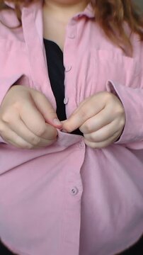 Close-up of a plus-size woman's hands unbuttoning a pink shirt, revealing a black top. Vertical video for body-positive casual fashion retail, daily routine, and apparel marketing ads.