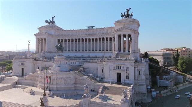 Aerial drone shot in Rome at Piazza Venezia showcasing the magnificent Altare della Patria monument with its white marble columns, bronze quadriga statues, and Italian flag under clear blue sky.