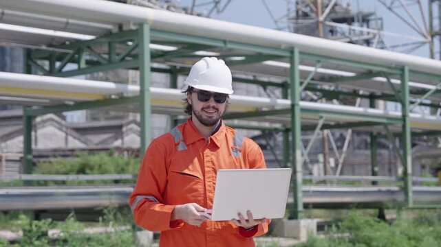 Smiling engineer confidently presents laptop technology before colossal industrial pipe structure during bright, sunny workday emphasizing operational progress and modern solutions.