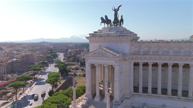 Aerial drone shot in Rome at Piazza Venezia showcasing the majestic Vittoriano monument with its bronze quadriga statue. The Colosseum and Roman Forum ruins stretch across the historic cityscape.