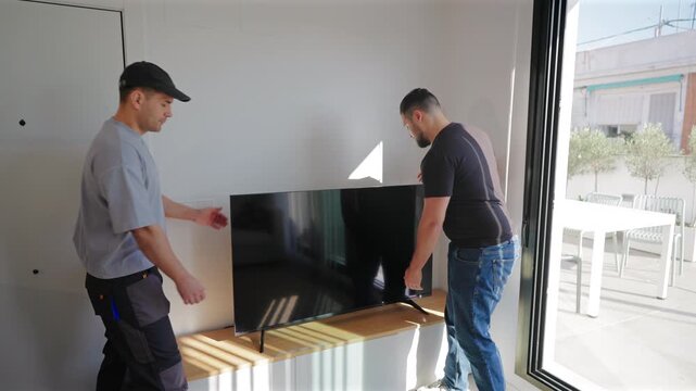 Two professional movers carrying large flat screen television at home during relocation service. Men carefully position TV on wooden stand in bright modern living room with natural light from window.