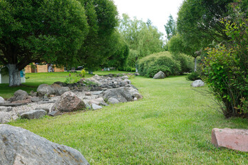 Fototapeta premium Beautiful rock garden with decorative natural stones on green grassy lawn. Professional landscaping design featuring dry creek bed with large boulders surrounded by lush green trees. Scenic