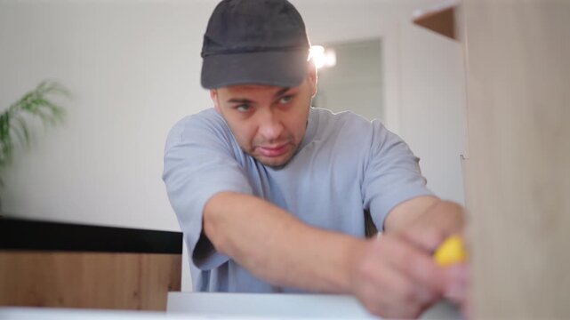 Young man wearing cap uses screwdriver from toolbox to perform maintenance in kitchen of home for repair. Worker focuses on assembling furniture with precision and concentration indoors.