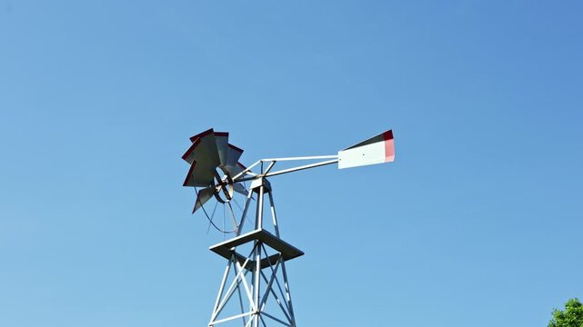 A windmill turns slowly in the sunlight with its blades reaching for the sky. The open space around highlights the rural setting and shows a clear blue sky above.
