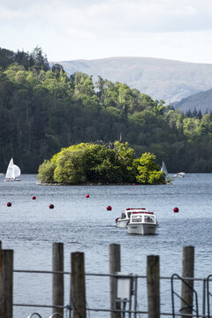 View of Windermere lake featuring small boats on the water and a lush green island with wooded hills in the background Windermere, England, United Kingdom.