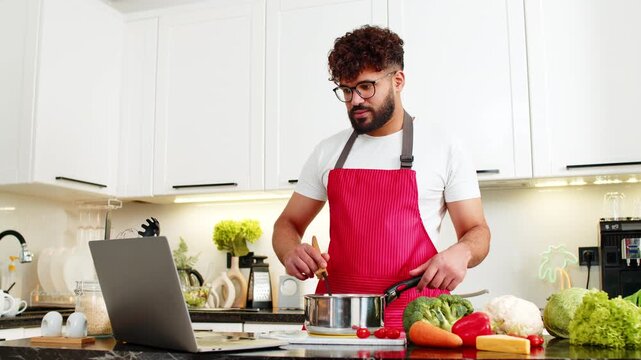 Arabian man stirring soup in pot on stove watching laptop video recipe tips in kitchen for healthy meal prep. Young guy focused on home cooking enjoying warm dinner for nutrition in apartment indoors.
