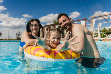 Obraz premium Young family enjoying time together in swimming pool, smiling and playing with inflatable ring under clear blue sky and bright sunlight