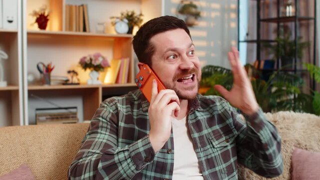 Young man on home sofa argues on smartphone with raised hand during tense call over bad offer. Adult guy on home sofa rejects false claim and explains point with anger while speaking in frustration.