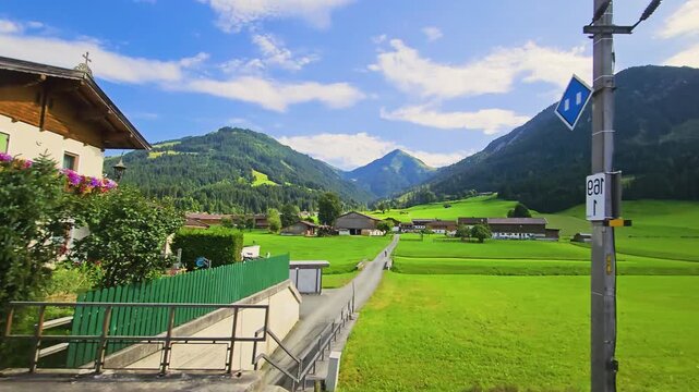Train travel scene with bright grass fields, village rooftops, forest slopes, and mountain peaks, filmed from a window while moving through the Alps in peaceful rural scenery.