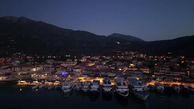 Aerial side-tracking drone shot along the illuminated waterfront of Nidri, Lefkada before sunrise. Ferries, excursion vessels, yachts and marina infrastructure line the harbor while warm city lights