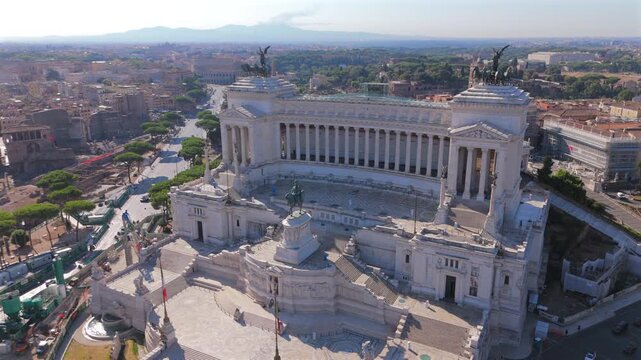 Aerial drone shot in Rome at Piazza Venezia featuring majestic Altare della Patria monument with its white marble columns and bronze quadriga statues overlooking the historic cityscape and Colosseum.