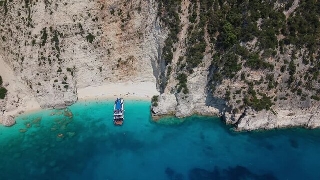Drone aerial view of a remote beach on Ithaca Island, Greece. Crystal-clear turquoise waters, white limestone cliffs and a tour boat anchored near the shore create a stunning Mediterranean summer dest