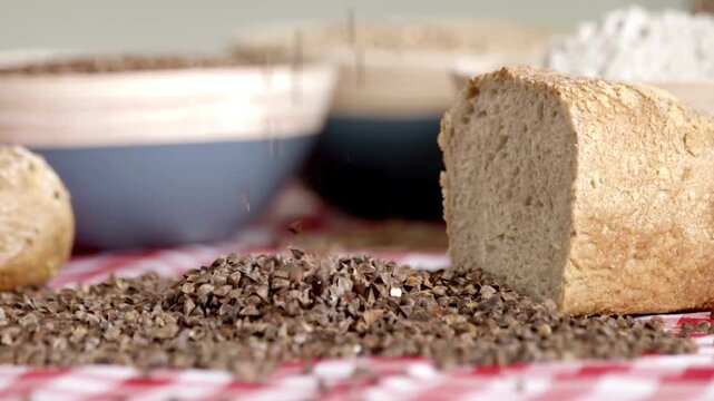 Buckwheat groats falling onto a pile next to a loaf of fresh bread on a checkered tablecloth