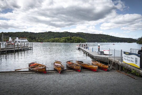 View of wooden rowboats on a slipway by the pier with people and a ferry on Lake Windermere under a cloudy sky Windermere, England, United Kingdom.