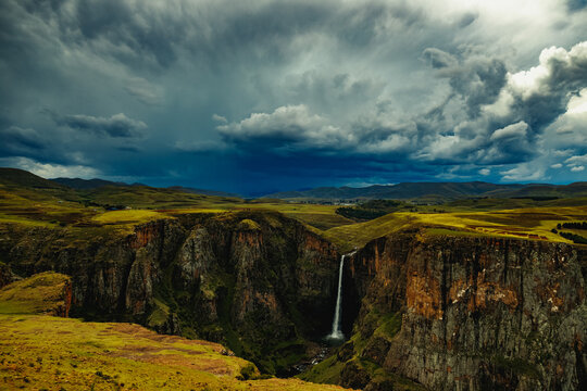 View of Maletsunyane Falls plunging into a deep rocky canyon under dramatic dark storm clouds and green rolling hills in Maseru, Lesotho.